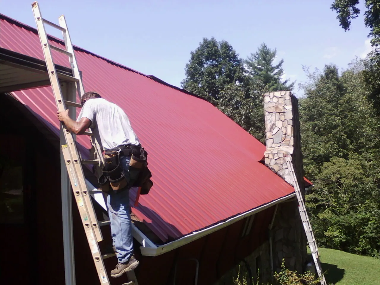 Roofer on a ladder beside a red metal roof and stone chimney — Soffit & Fascia Repair by Roofing Brothers in Blakely Island